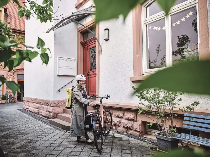 Eine selbstständige Pendlerin mit Regenmantel steht in urbaner Gegend neben ihrem Fahrrad