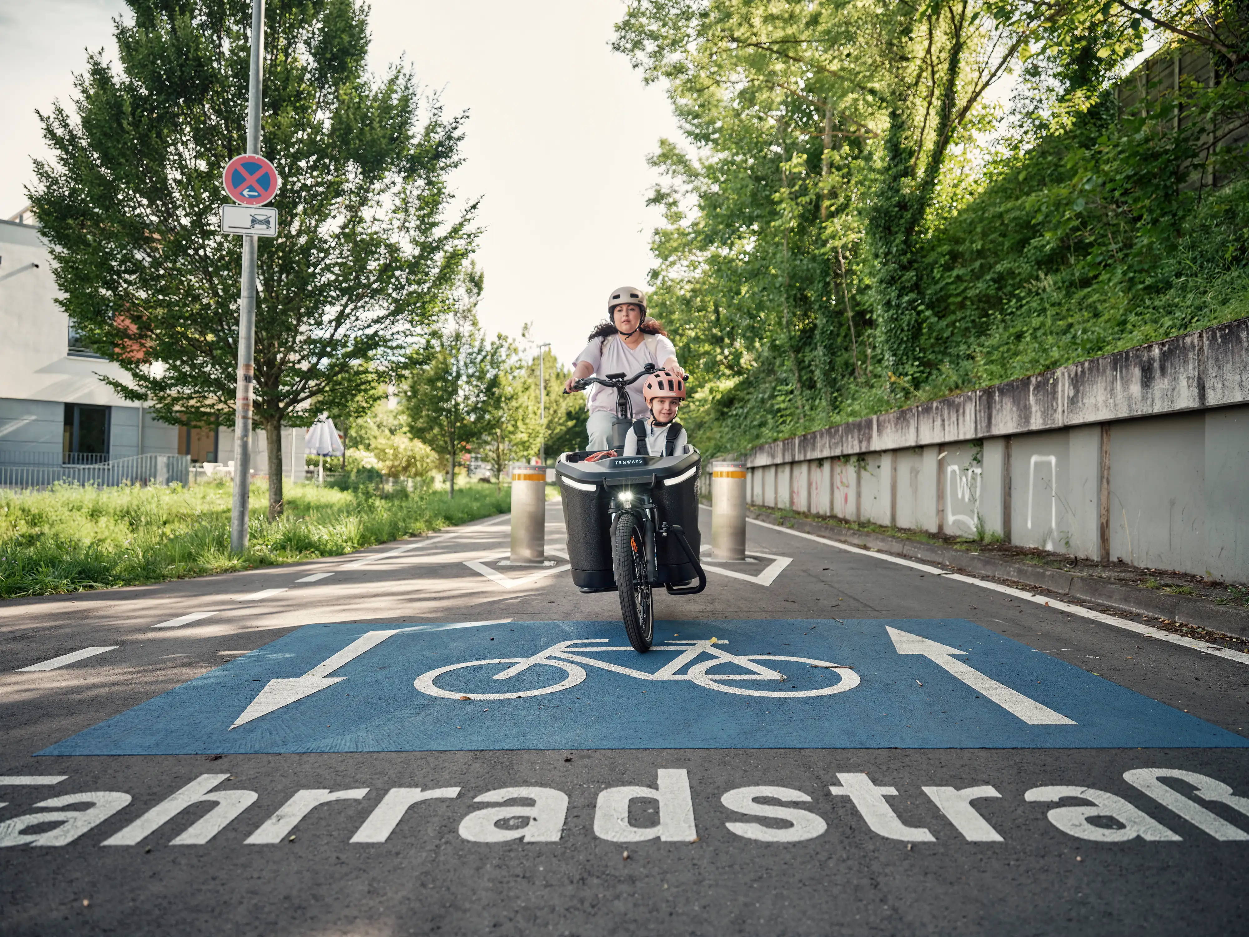 Mama mit Kind im Lastenrad fährt auf Fahrradstrasse zwischen Pollern hindurch